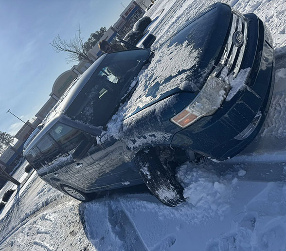 A blue Ford SUV stuck in snow in a parking lot with snow covering parts of the vehicle on a sunny winter day.