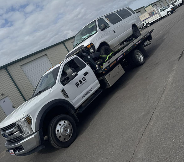White flatbed tow truck from G & G towing a white van in an industrial area under a cloudy sky.