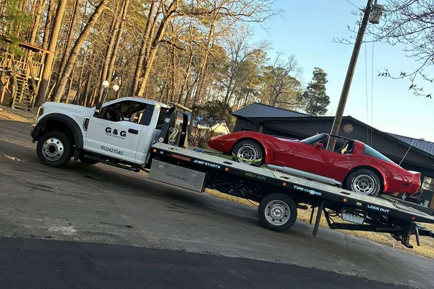 Flatbed tow truck from G&G transporting a red classic convertible car on a suburban street with trees and houses in the background.