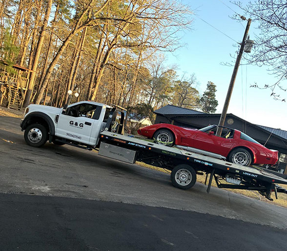 Flatbed tow truck from G&G transporting a red classic convertible car on a suburban street with trees and houses in the background.