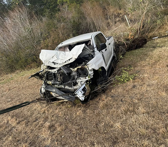 White pickup truck with severe front-end damage being towed from a grassy roadside area with trees in the background.